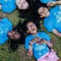 Four diverse girls laying in the grass smiling wearing Girls on the Run program shirts.